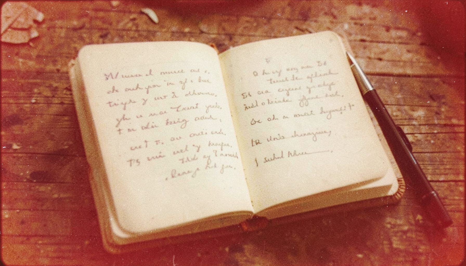 A grainy analog film photograph of an open notebook with handwritten text and a pen on a rustic wooden surface, in warm brown tones
