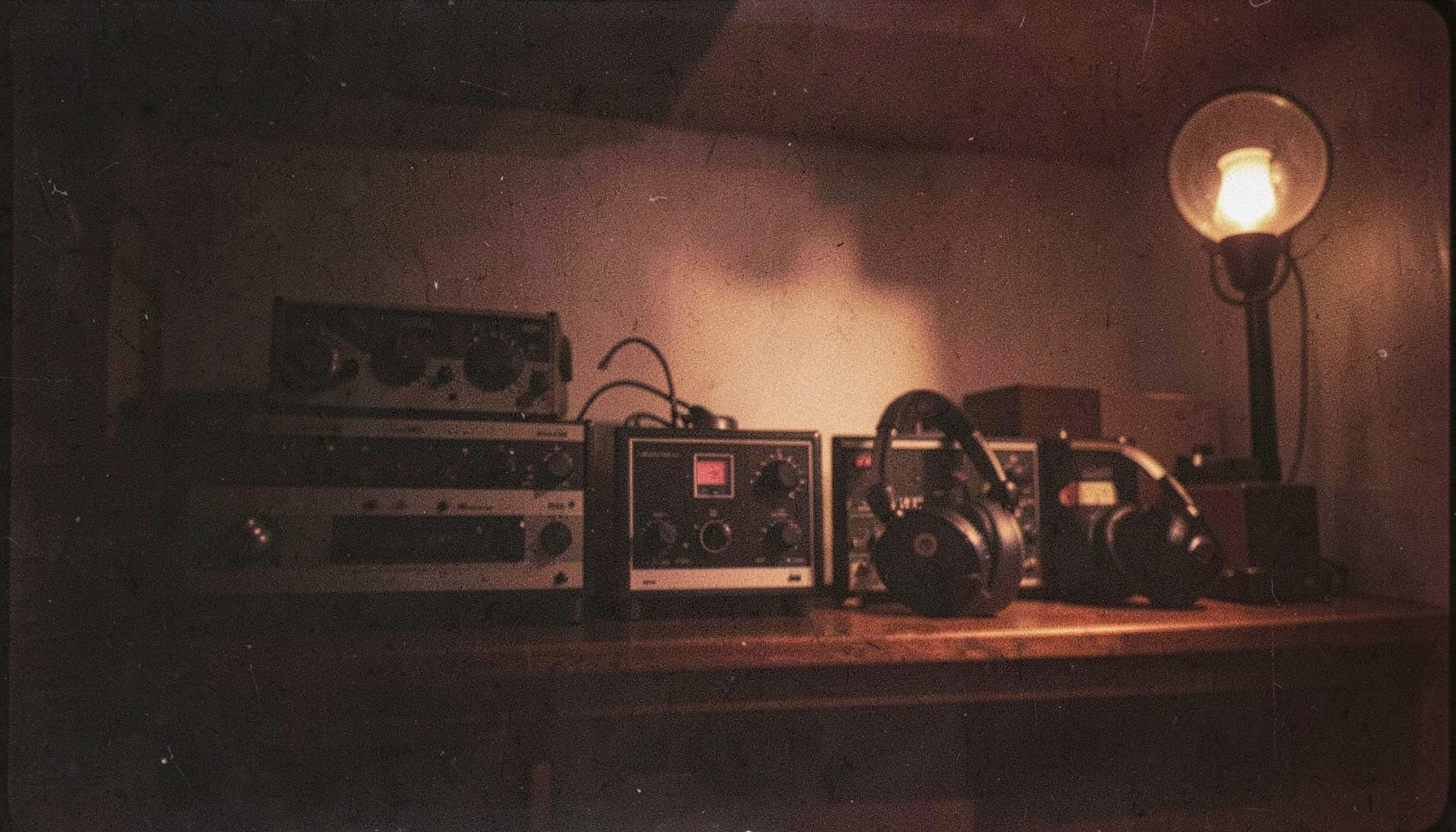 A grainy analog film photograph of vintage audio equipment and headphones on a dark wooden shelf, lit by a single warm lamp
