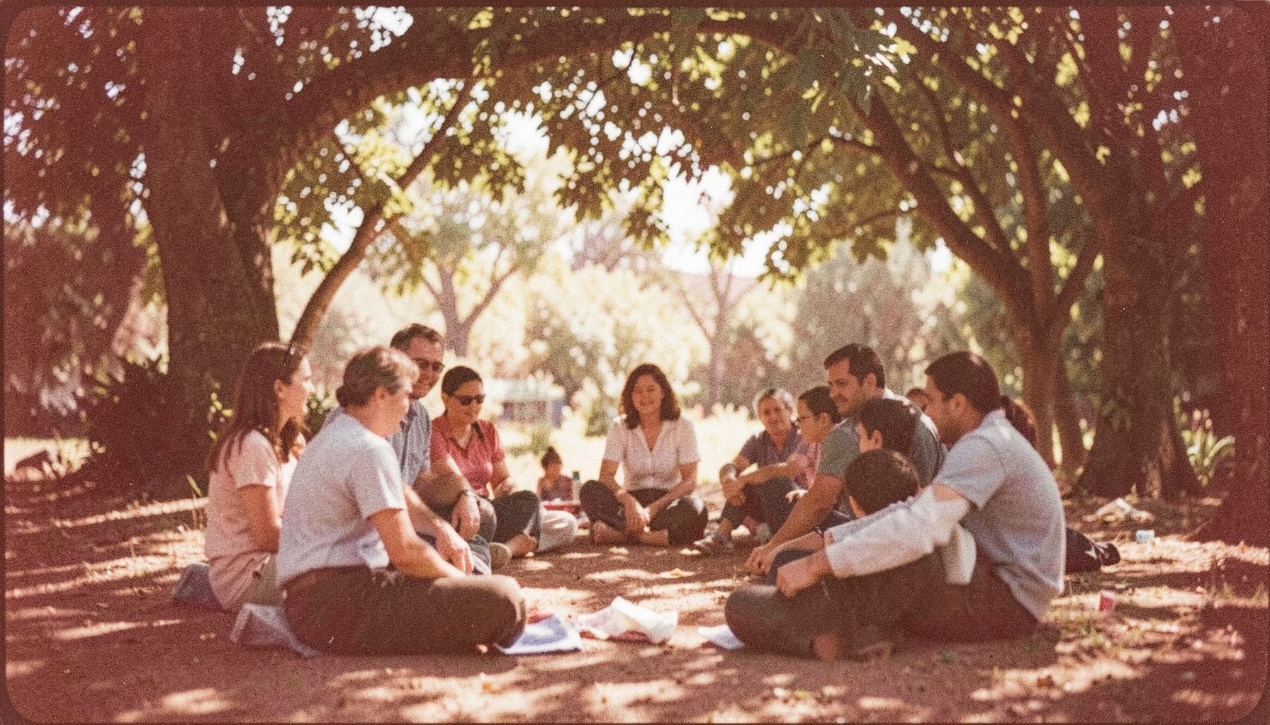 A grainy analog film photograph of people seated in a loose circle outdoors under dappled tree shade, with warm earthy browns and greens